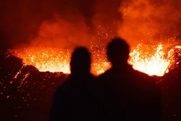 Gece vakti kızgın lav püskürten bir yanardağ kraterini uzaktan izleyen iki kişinin silueti