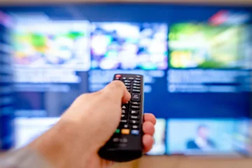 Elinde televizyon kumandası tutan bir kişi, arkada bulanık bir dizi seçim ekranı