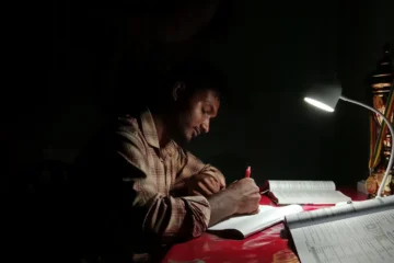 Karanlık odada masa lambasının ışığında defterine not alarak ders çalışan genç